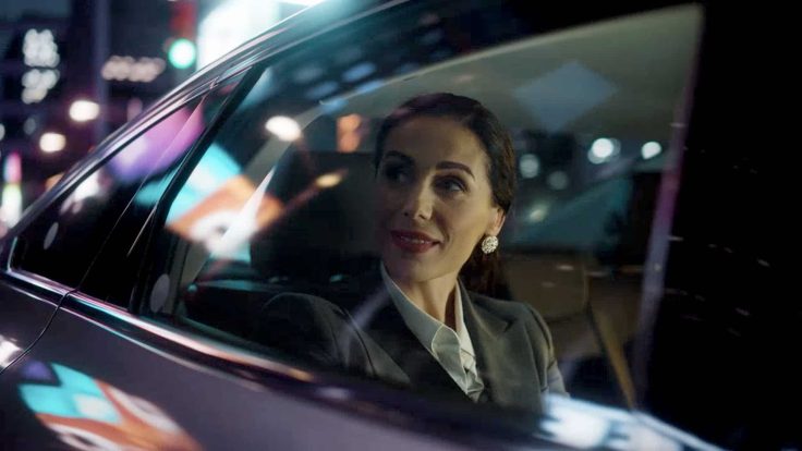 A woman sitting in a car looking out the window with a city reflected on the car's surface.