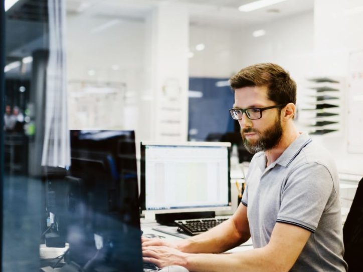 Man working at a desk in a modern office environment, focused on his computer screen with reflections visible on the glass wall beside him.