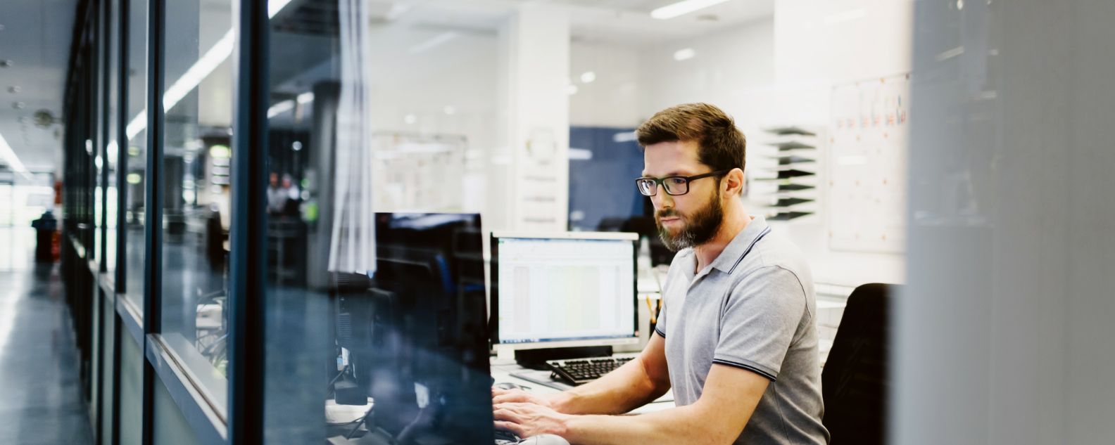 Man working at a desk in a modern office environment, focused on his computer screen with reflections visible on the glass wall beside him.